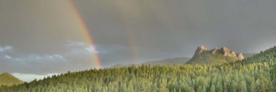 A tent sits in an open field in front of a forest of trees and a double rainbow